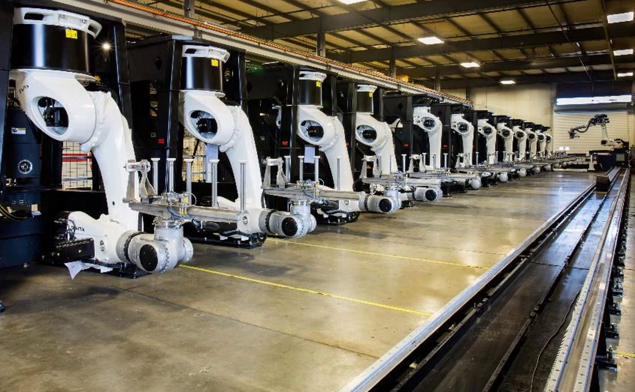 A line of robots stands ready to position various carbon fiber parts for non-destructive inspection by the robot in the background, which is equipped with an ultrasonic test head and sits on a track on which it can move up and down the line. Source: ATI Automation