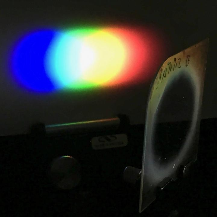 This photo shows a one-inch diameter Bragg polarization grating diffracts white light from an LED flashlight onto a screen placed nearby. Source: Michael Escuti, NC State University